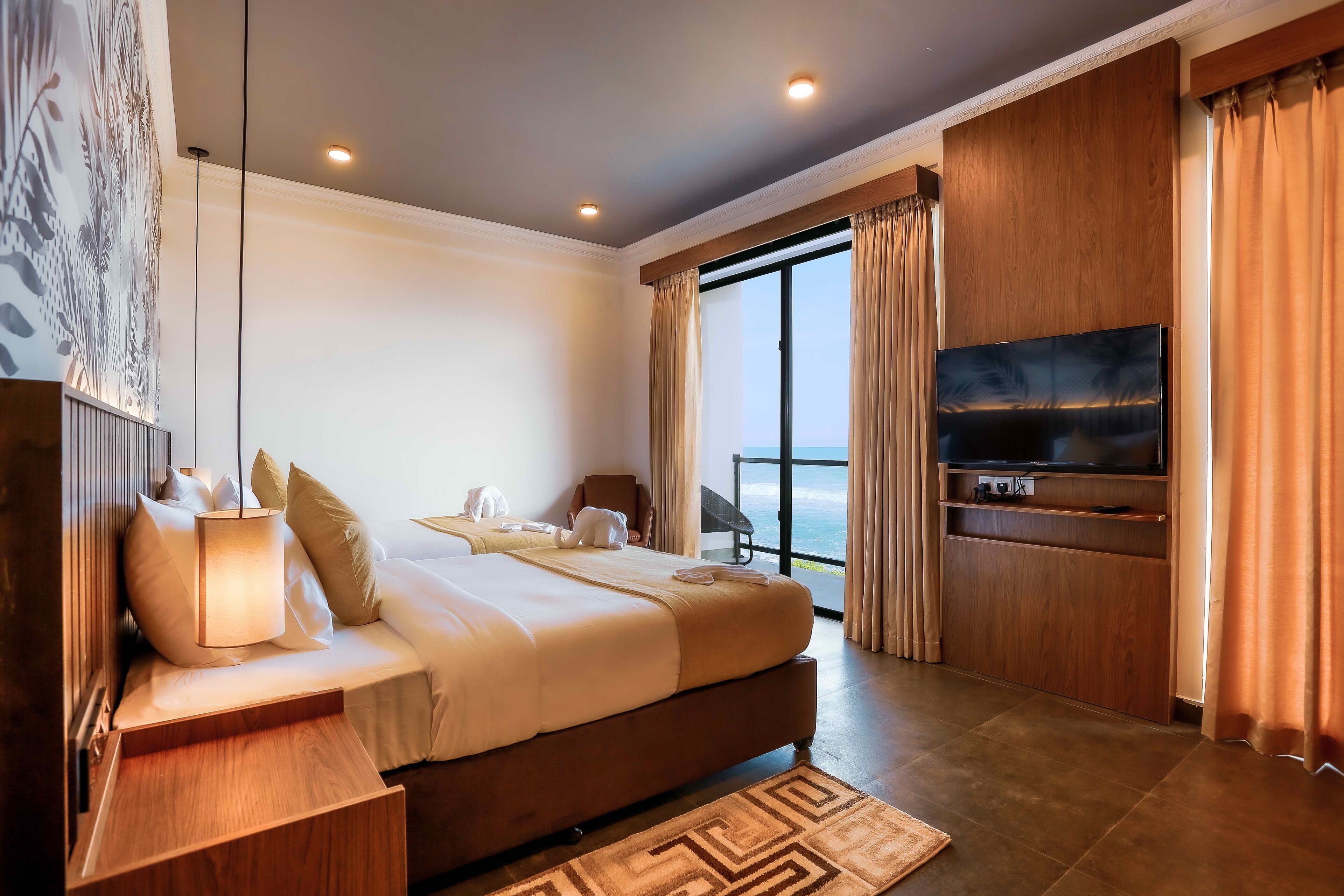 A modern hotel room with a large bed, a TV, a balcony with curtains, and a view of the ocean.