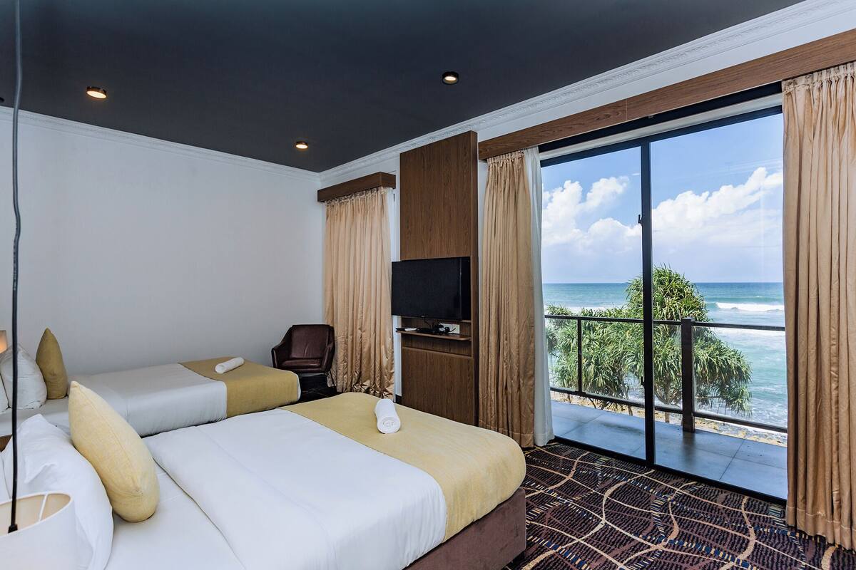 A hotel room with two beds, a TV, and a balcony with a view of the ocean.