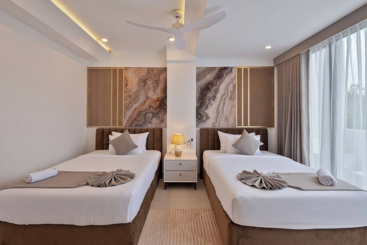 A hotel room with two beds, a ceiling fan, a nightstand with a lamp, and a large window with curtains.