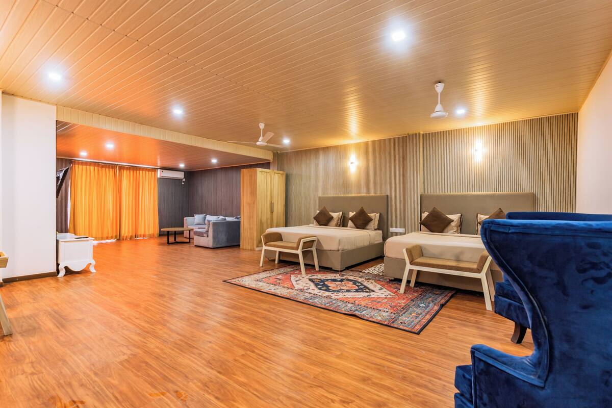 A modern hotel room with a wooden ceiling, two beds, a blue armchair, a small table, and a patterned rug.