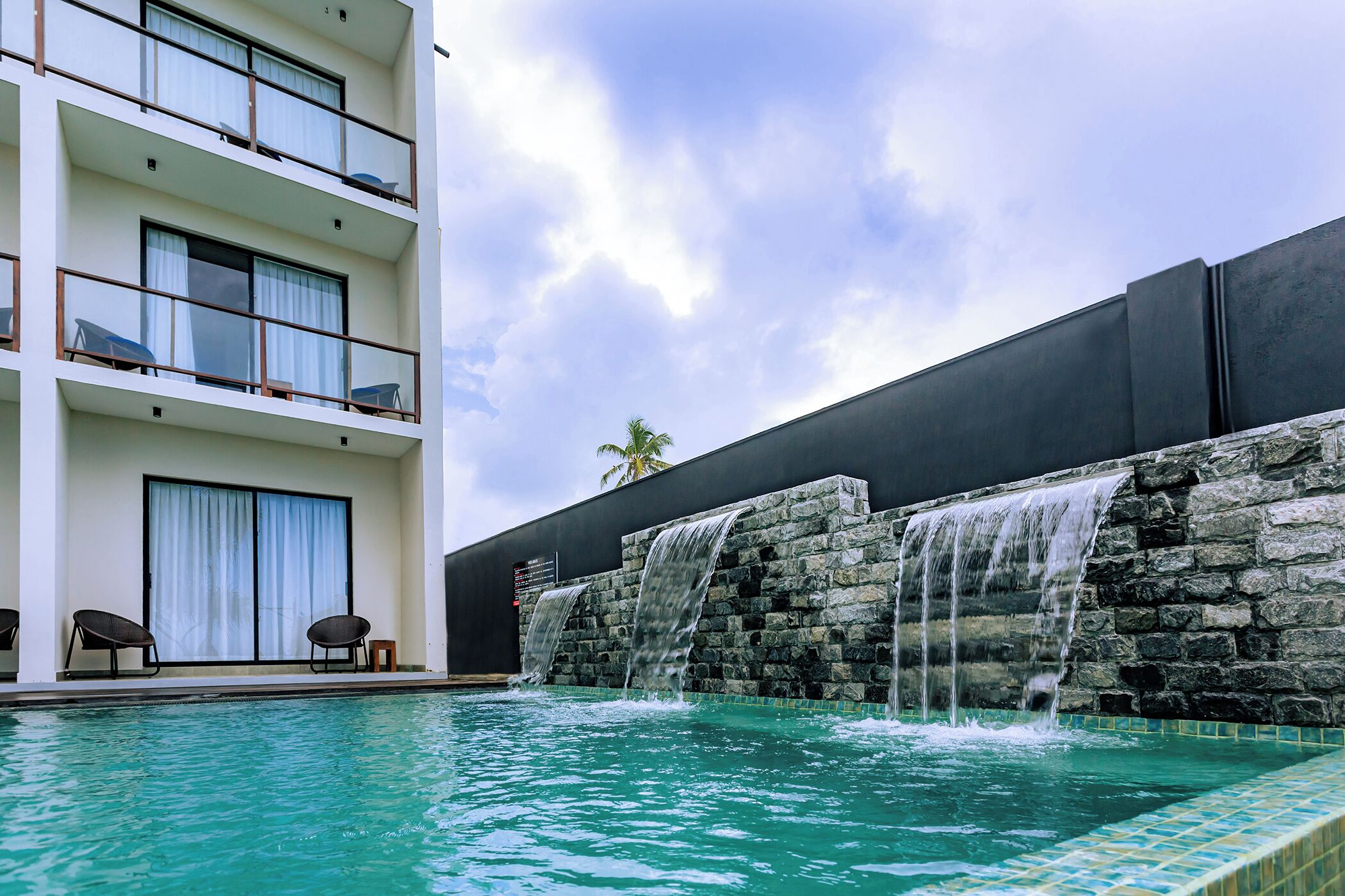 A swimming pool with a waterfall feature, a multi-story building with balconies, and a palm tree in the background.