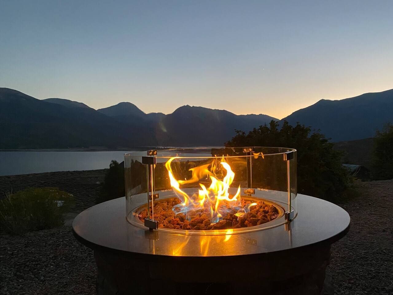 Ein kreisförmiger Feuerkorb im Freien mit Flammen, umgeben von einer klaren Glaseinfassung, vor einer Bergkulisse.