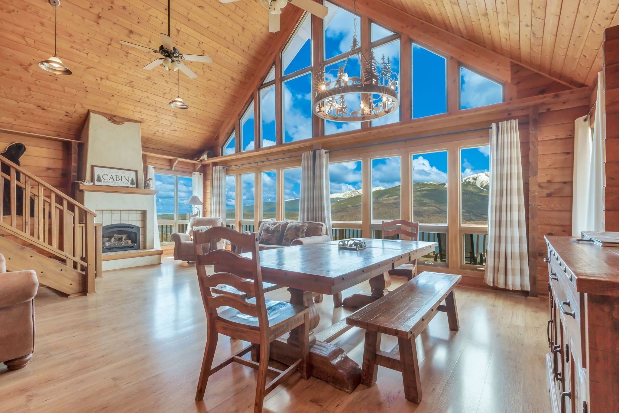 Ein geräumiges Blockhaus-Interieur mit großem Esstisch, Holzsesseln, Kamin und großen Fenstern mit Blick auf die Berge.