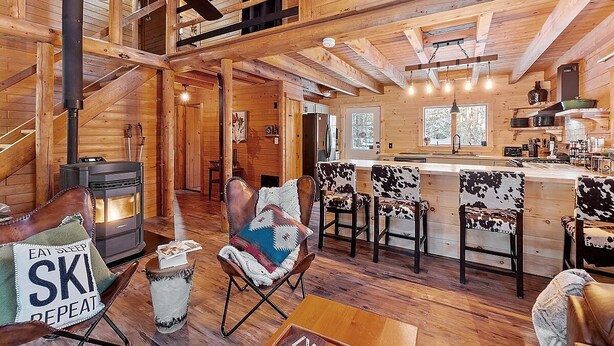 Un intérieur de chalet chaleureux avec un espace salon central, une cuisine dotée d’un bar et un poêle à bois.