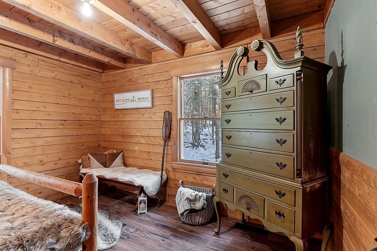 A cozy cabin room with wooden walls, a bed with a fur blanket, a dresser, and a window with a snowy view.