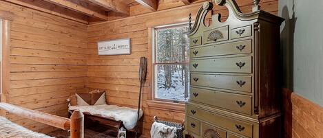 A cozy cabin room with wooden walls, a bed with a fur blanket, a dresser, and a window with a snowy view.