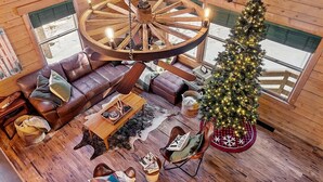 Un intérieur de chalet chaleureux avec un grand lustre en forme de roue en bois, un sapin de Noël, des canapés en cuir et un plancher en bois rustique.