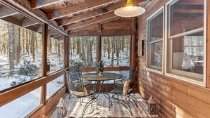 Une cabane en bois dotée d’un coin repas avec une table ronde et des chaises, ainsi que de grandes fenêtres offrant une vue sur une forêt enneigée.