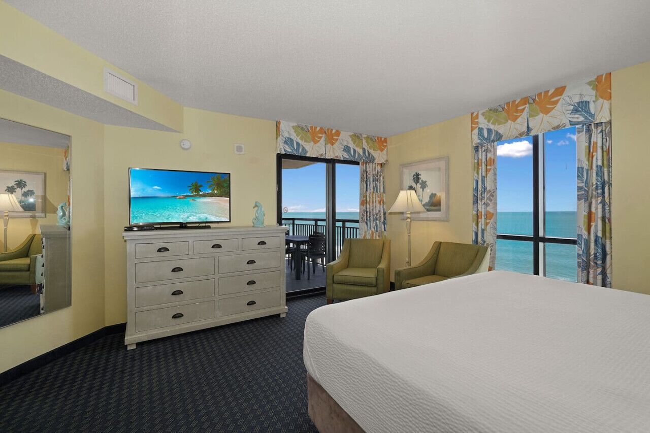 A hotel room with a bed, a television, a dresser, and a view of the ocean.