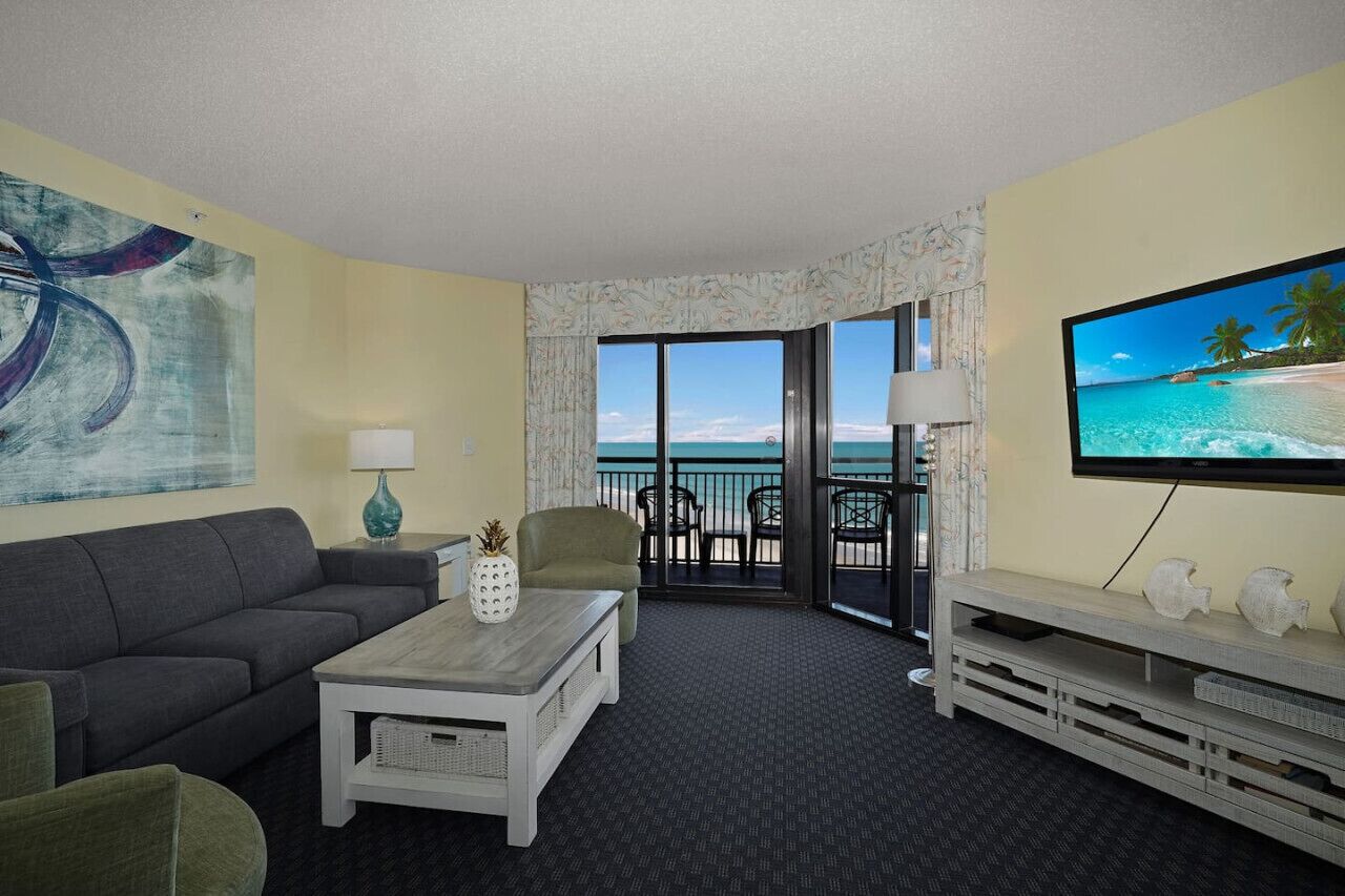 A hotel room with a sofa, coffee table, TV, and a balcony with a view of the ocean.