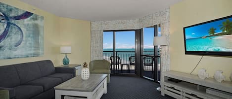 A hotel room with a sofa, coffee table, TV, and a balcony with a view of the ocean.