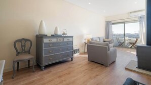 Een moderne woonkamer met een grijze bank, een houten vloer, een grijze dressoir en uitzicht op de stad door grote ramen.