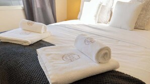 Een hotelkamer met een net opgemaakt bed, twee opgerolde handdoeken met een logo, een donker geweven vloerkleed en een raam met gordijnen.