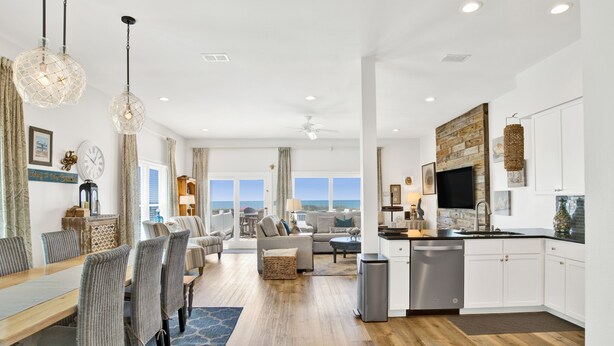 A spacious living area with a dining table, sofas, and a kitchen with white cabinets and a large window. - Gulf-Front Duplex | Pet Friendly | Pools and Pier Access (Gulf Shores)