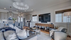 A modern living room with a sofa, coffee table, and a TV. There is a kitchen area with cabinets and a dining table.