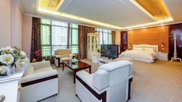 A hotel room with a large bed, a TV, a sofa, and a desk. There is a window with curtains and a vase of flowers on the side table. - Shengjing Hotel (Fuyuan)