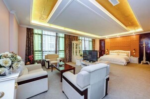 Habitación de hotel con una cama grande, televisión, sofá y escritorio. Tiene una ventana con cortinas y un jarrón con flores sobre la mesita de noche.