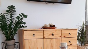 Uma mesa de madeira com uma garrafa de vinho, copos e fruta. Uma televisão está fixada na parede acima.