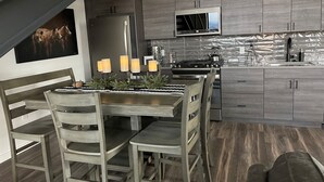 A modern kitchen with grey cabinetry, a stainless steel refrigerator, a microwave, and a dining area with a table and chairs. - MT Luxury! (Bozeman)
