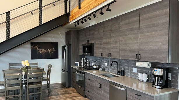 A modern kitchen with a wooden staircase, a dining area with a table and chairs, and a kitchen island with a sink and coffee maker. - MT Luxury! (Bozeman)