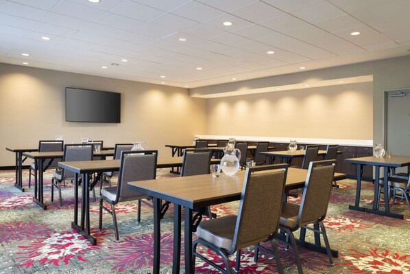 A conference room with multiple tables and chairs, a large screen on the wall, and a carpeted floor.