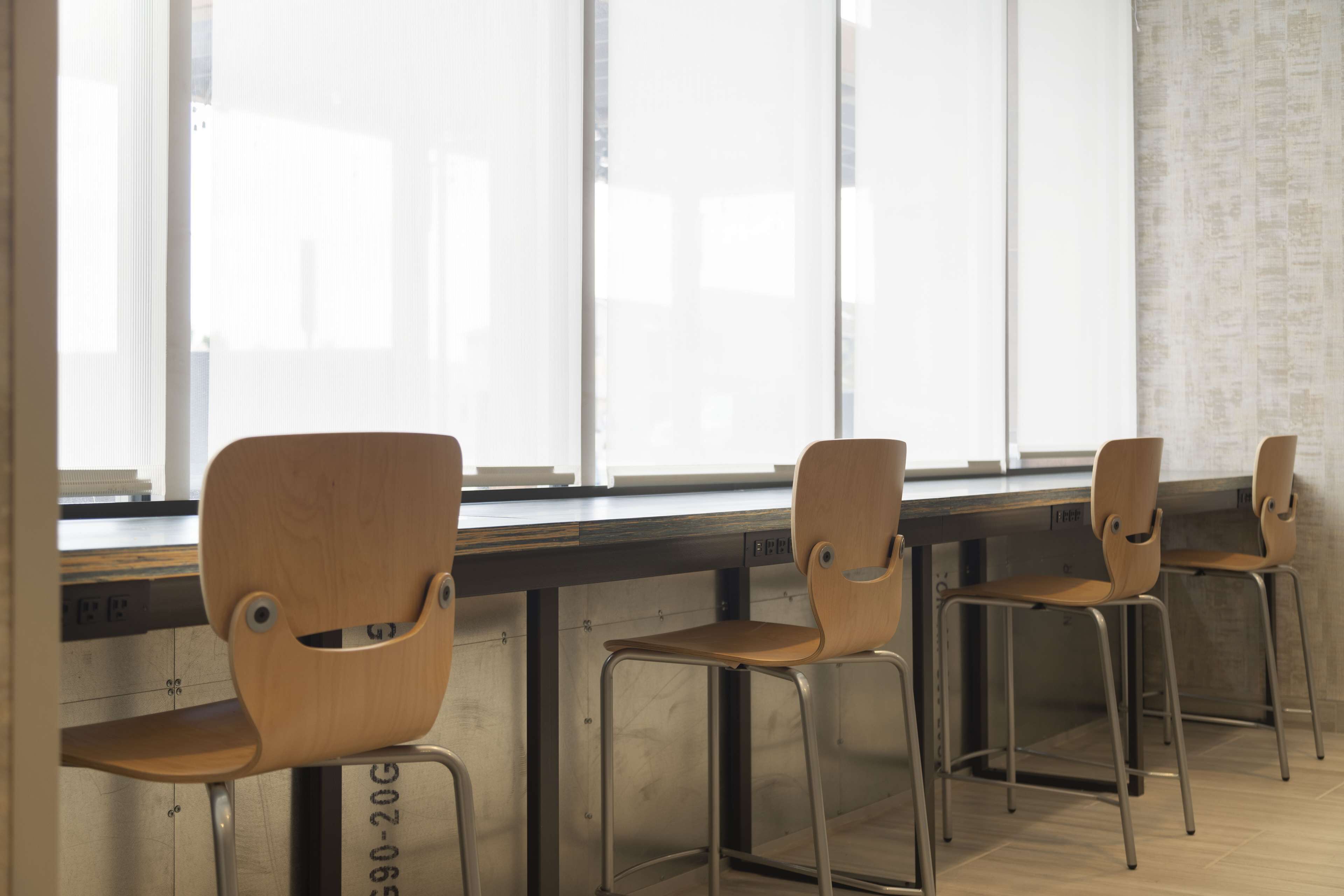 A row of wooden chairs with metal frames, positioned in front of a window with a view of buildings.