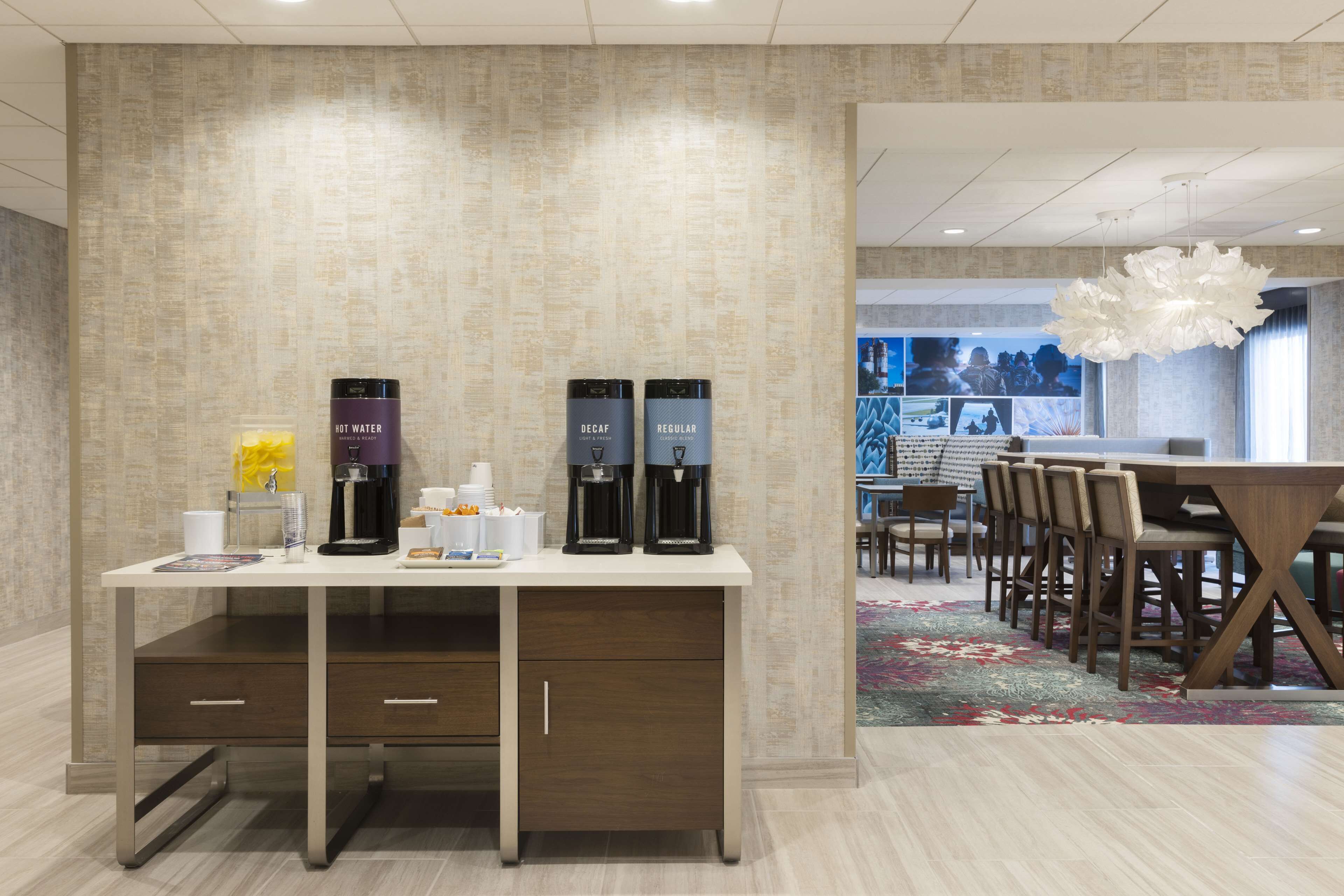 A hotel reception area with a reception desk, coffee machines, and a seating area with chairs and a table.