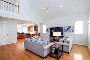 A spacious living room with a grey sofa, a coffee table, and a TV. There is a kitchen in the background with wooden cabinets and a dining area. - All Seasons Villa (Captiva)