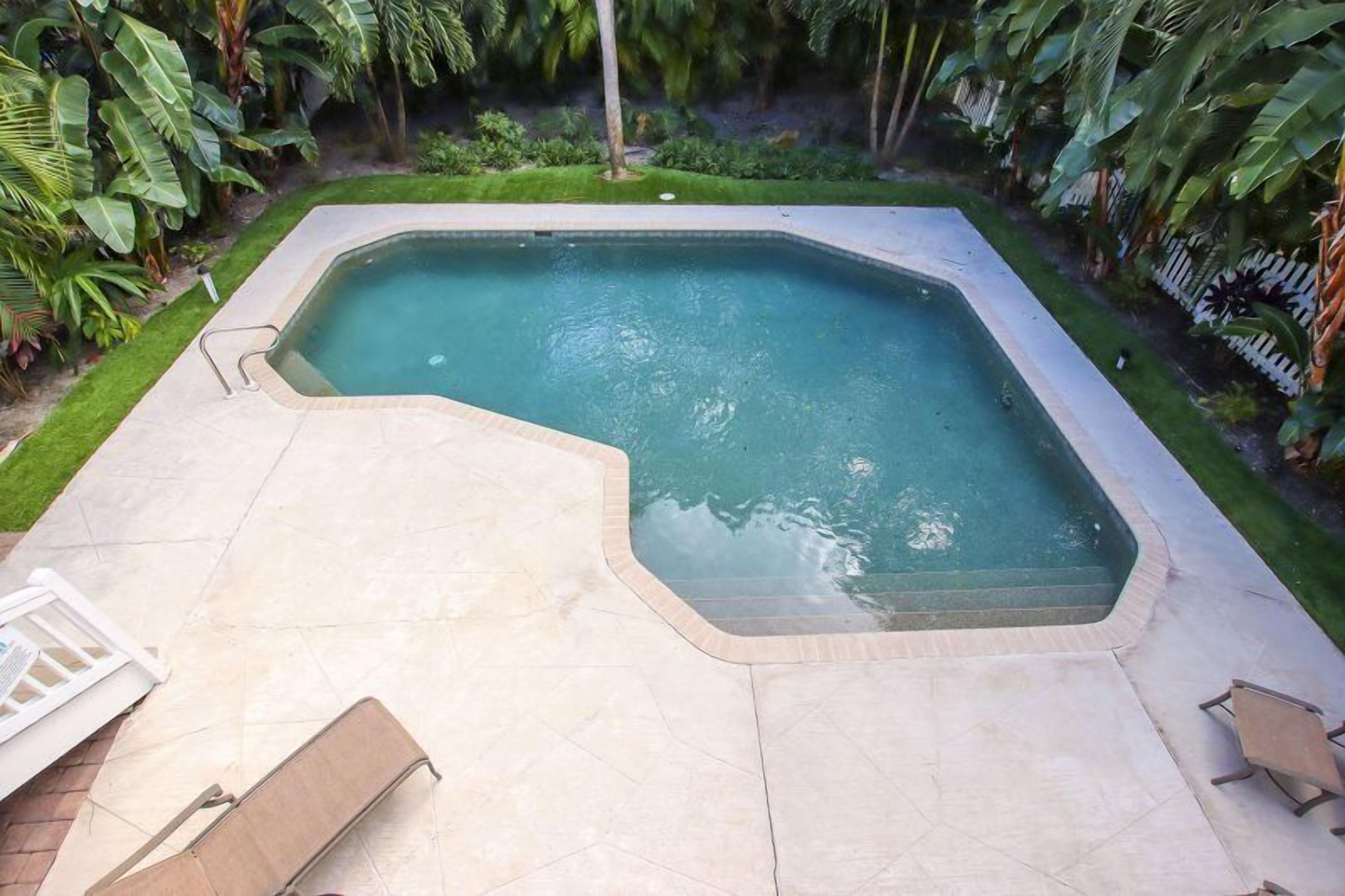 A rectangular swimming pool with a tiled deck, surrounded by lush greenery and a white fence.