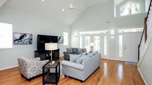 A spacious living room with hardwood flooring, a grey sofa, a patterned armchair, a TV, and a ceiling fan. - All Seasons Villa (Captiva)