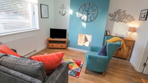 A living room with a blue accent wall, a large clock, a television, and a cozy seating area with a colorful rug. - Sea Glass (Filey)