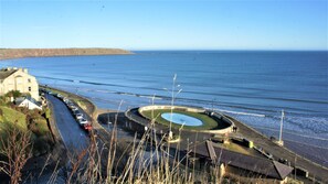 A coastal road with a roundabout, a beach, and a building with parked cars.