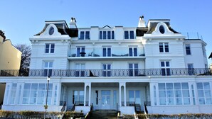 A large, white, multi-story building with a symmetrical facade, multiple balconies with ornate black railings, and a central entrance with steps leading up to it. - Sea Glass (Filey)
