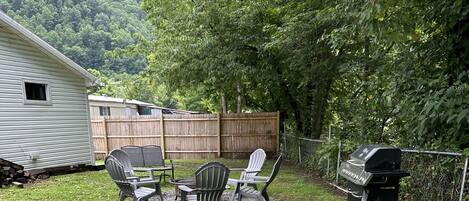 A wooden picnic table with chairs, a grill, and a fenced yard with trees.
