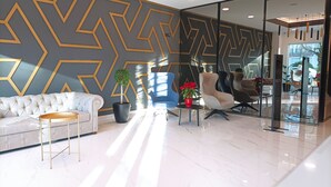 A modern hotel lobby with a geometric wall design, a tufted sofa, a round coffee table, and a potted plant. - Hotel Lux Melide (Melide)