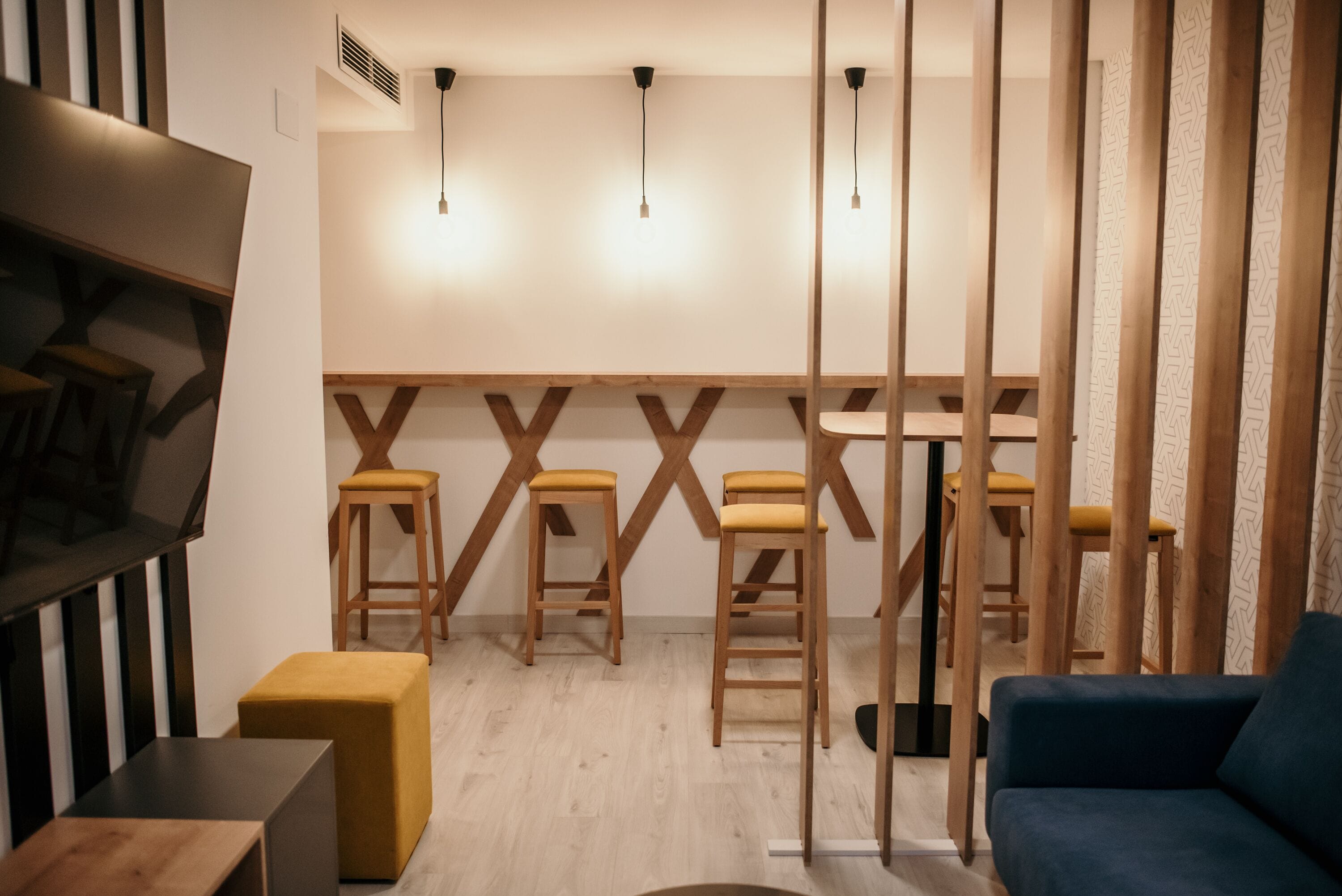 A modern interior with a bar area, wooden stools, a blue sofa, and a TV mounted on the wall.