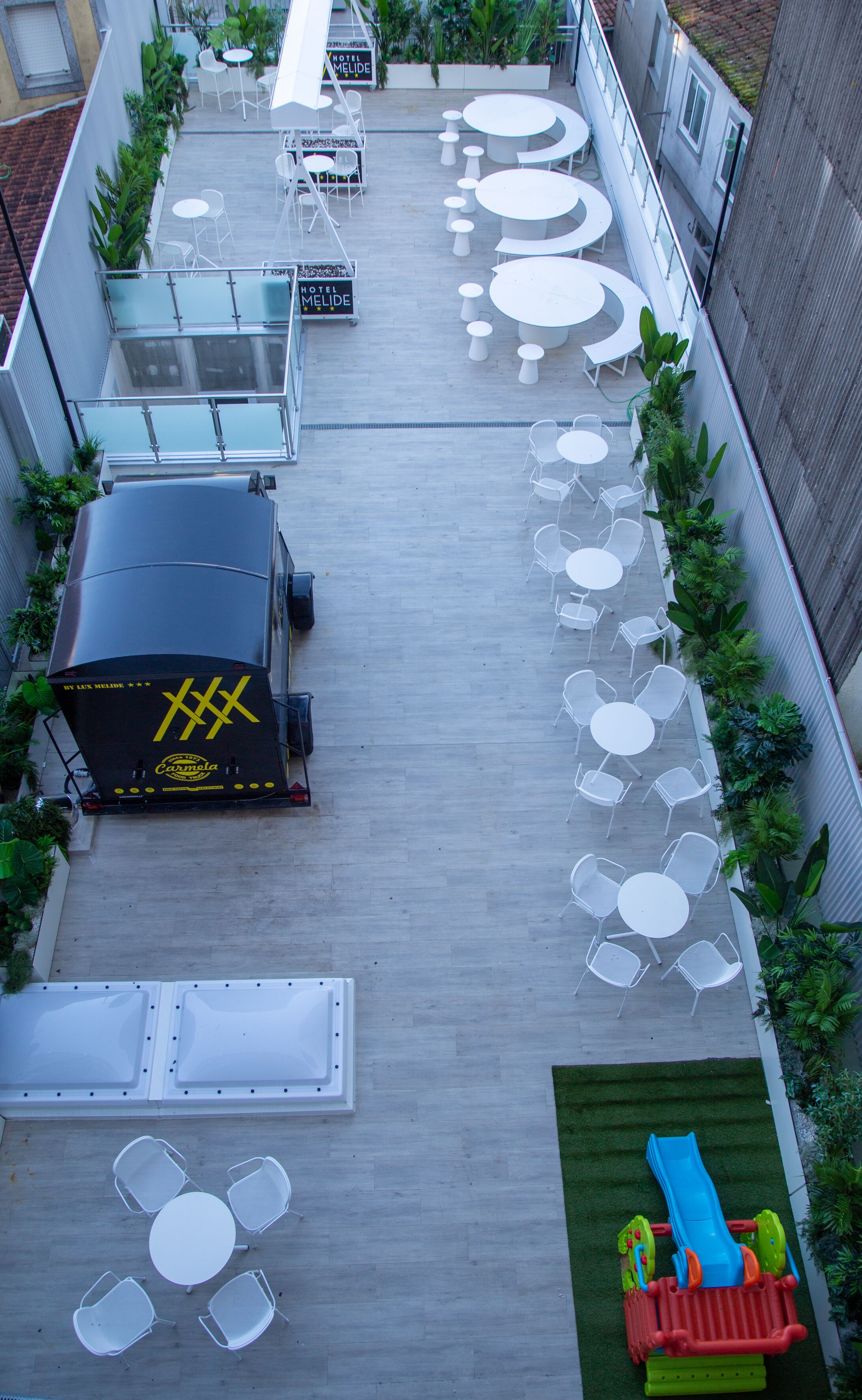 A rooftop terrace with multiple white round tables and chairs, a small play area with a slide, and a large black truck with yellow lettering.