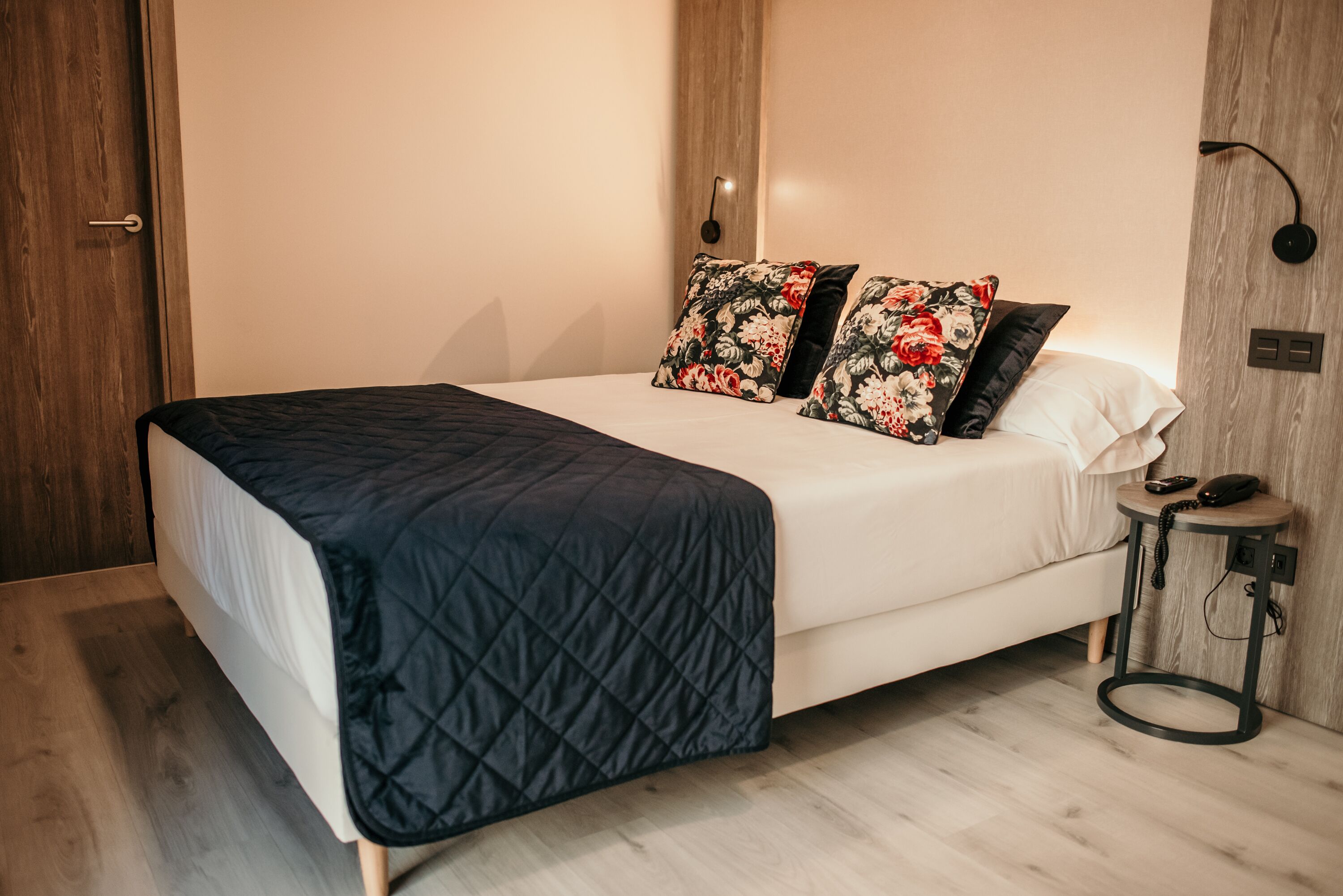 Una cama bien hecha con cubrecama acolchada, dos almohadas estampadas y una mesita de noche con un teléfono y una lámpara.