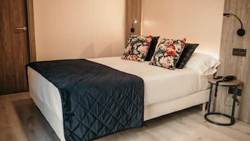 Una cama bien hecha con cubrecama acolchada, dos almohadas estampadas y una mesita de noche con un teléfono y una lámpara.