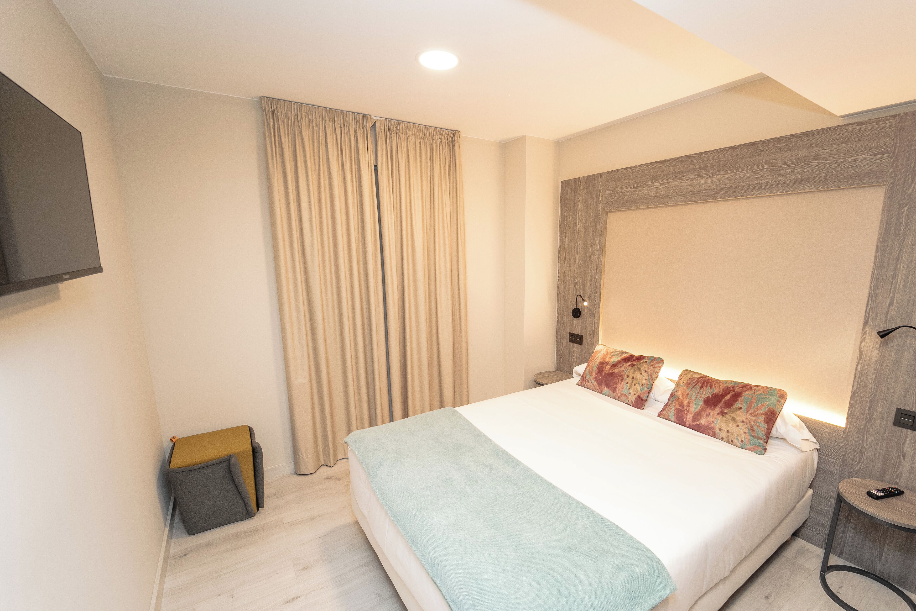 Ein modernes Hotelzimmer mit einem großen Bett, einem wandmontierten Flachbildfernseher, einem Nachttisch mit Lampe und einem kleinen Holzstuhl.