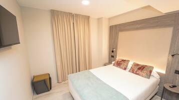 Una habitación de hotel moderna con una cama grande, un televisor de pantalla plana montado en la pared, una mesita de noche con lámpara y una pequeña silla de madera.