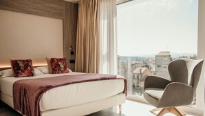 A modern bedroom with a large bed, a chair, and a city view through a large window. - Hotel Lux Melide (Melide)