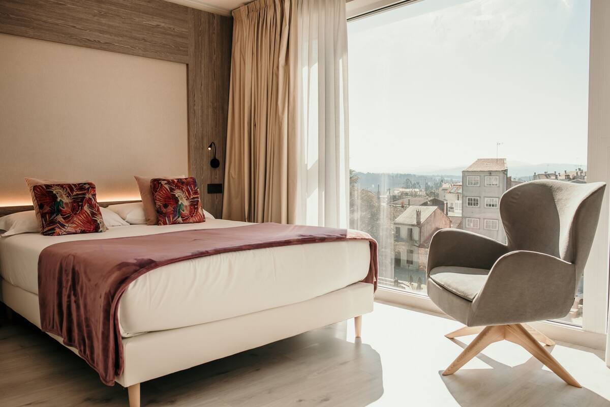 Ein modernes Schlafzimmer mit einem großen Bett, einem Sessel und Blick auf die Stadt durch ein großes Fenster.