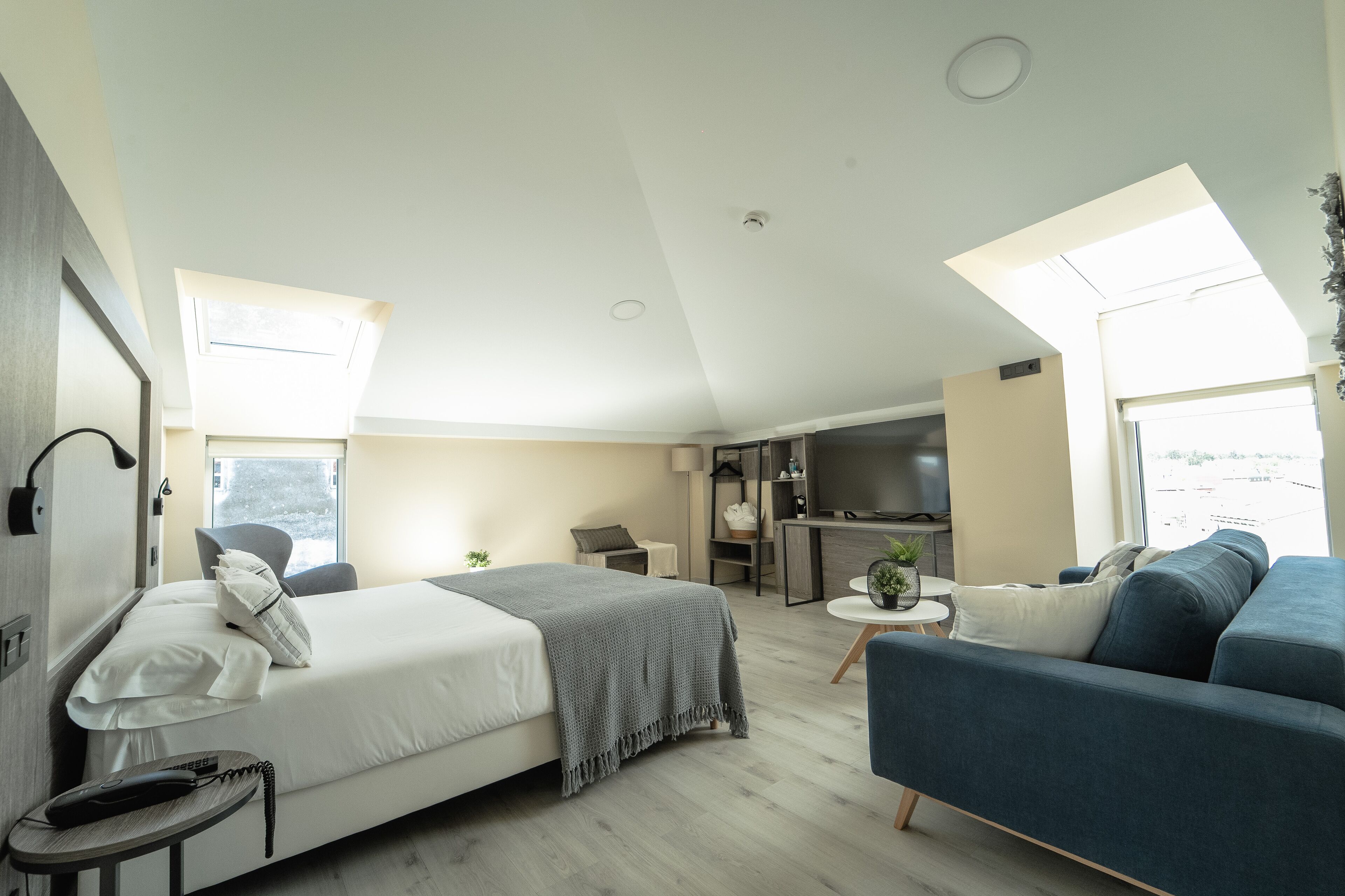 A modern bedroom with a bed, bedside table, and a desk with a chair. There is a sofa and a TV in the living area.