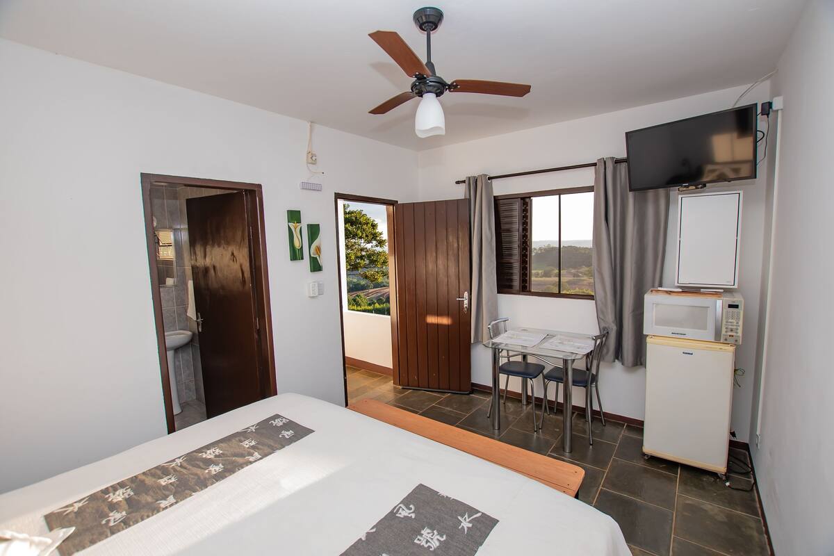 A hotel room with a bed, a ceiling fan, a television, a small refrigerator, a dining table with chairs, and a view of the outdoors.
