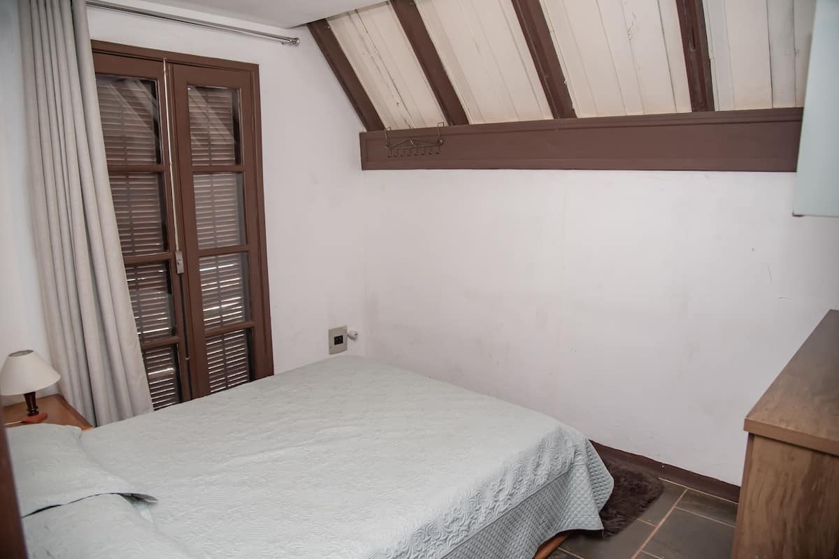 A bedroom with a bed, a window with blinds, a lamp, and a ceiling with wooden beams.