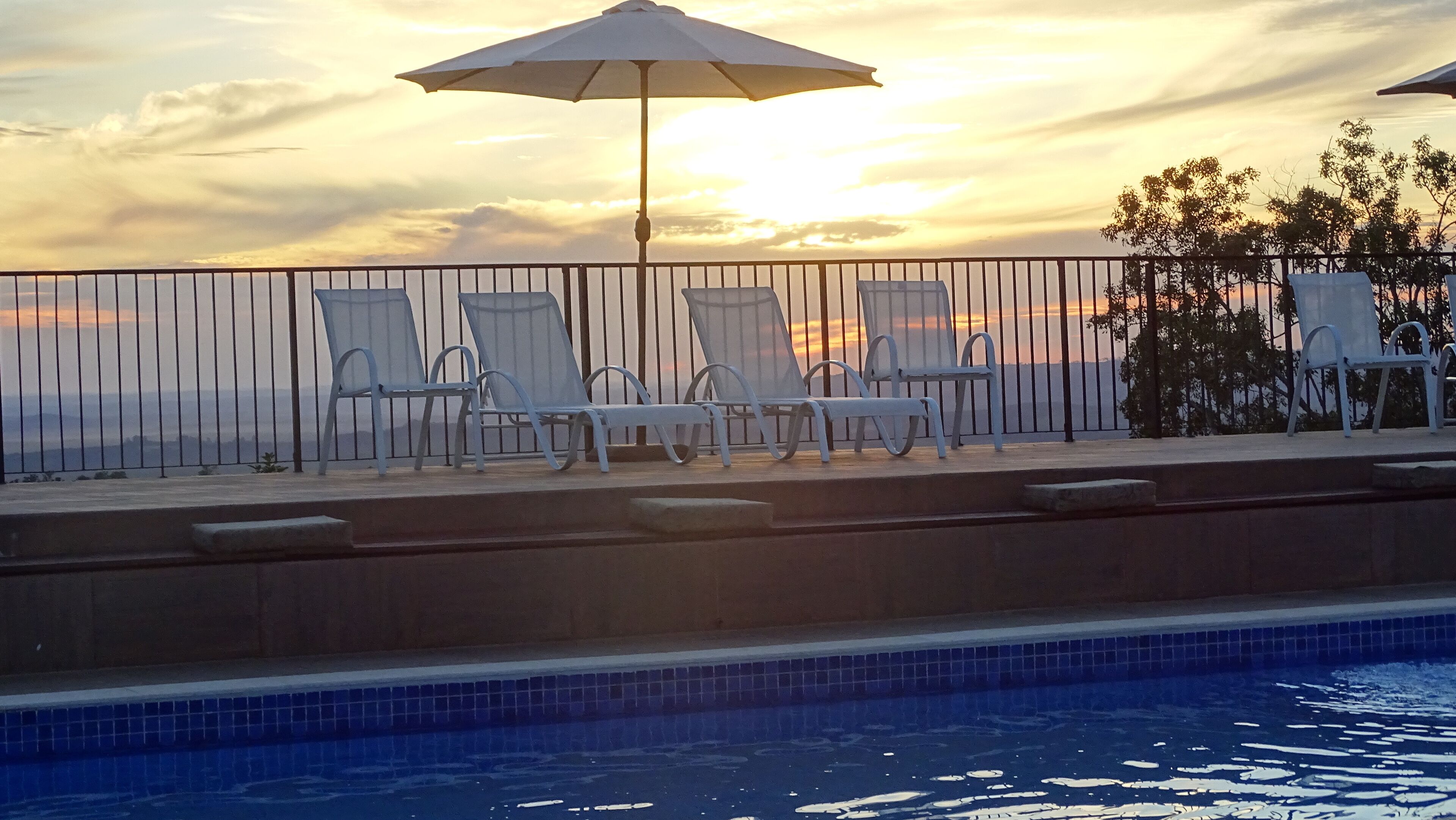 Un espace aménagé au bord de la piscine, avec des chaises longues et un parasol, offrant une vue sur une étendue d’eau au coucher du soleil.