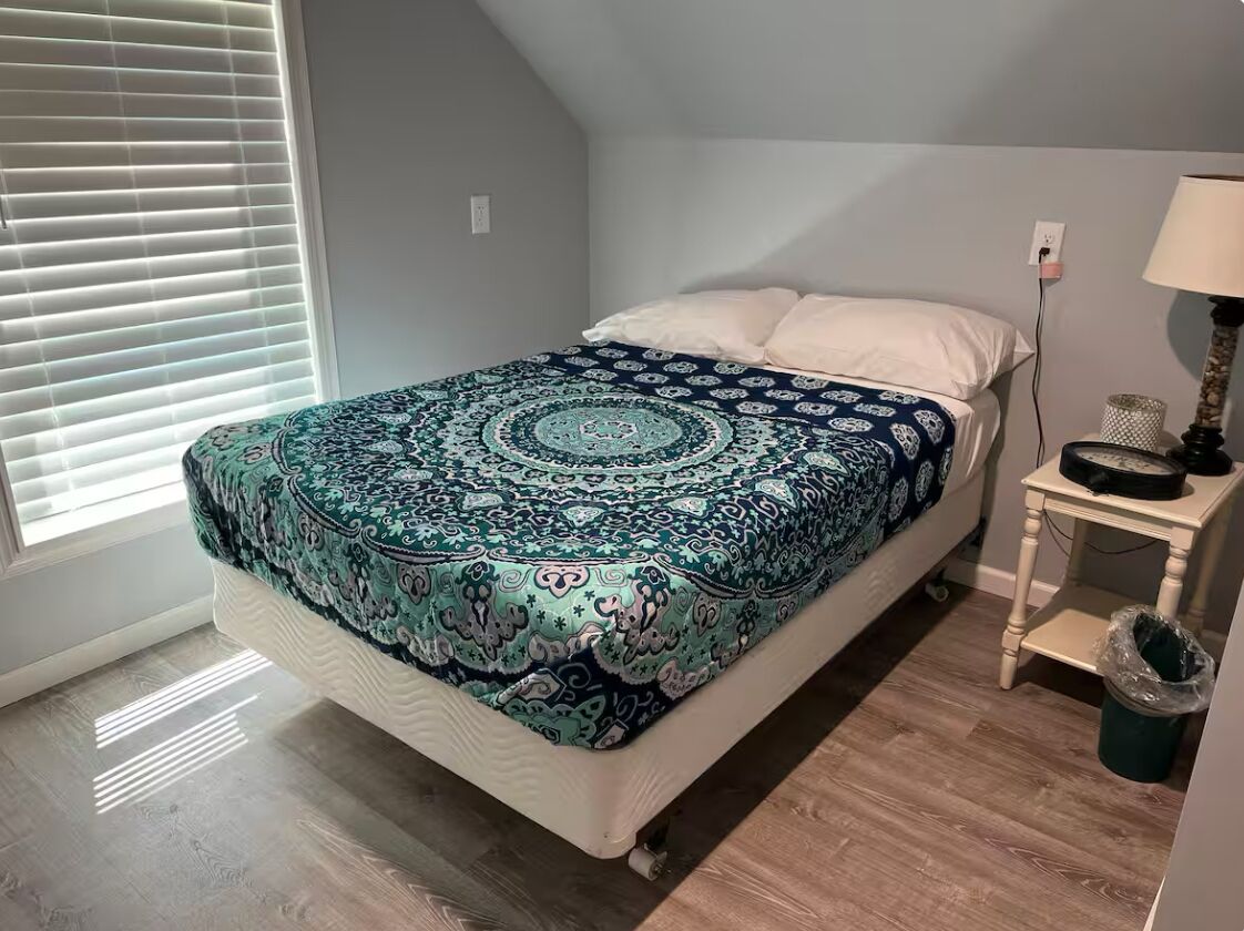 A bedroom with a bed, a bedside table, a lamp, and a window with blinds.