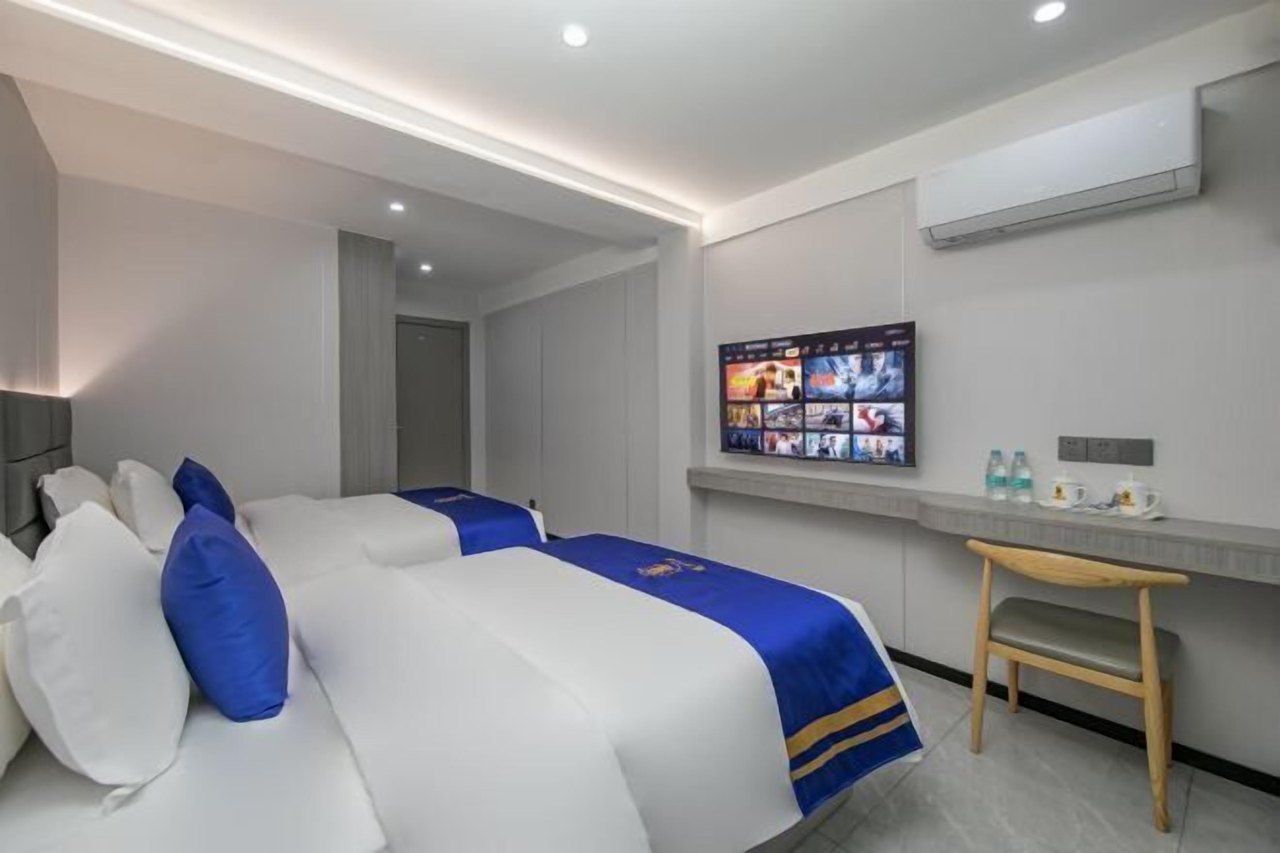 Quarto de hotel com duas camas, uma escrivaninha, uma cadeira e televisão na parede exibindo diversos canais.