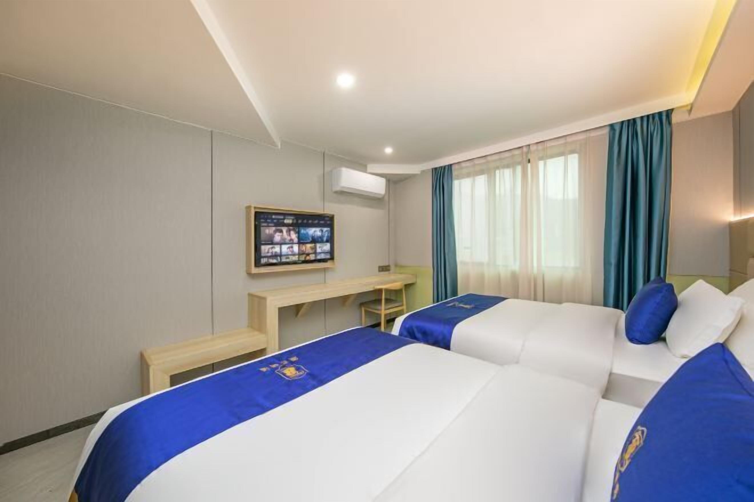 A hotel room with two beds, a desk, a television, and a window with curtains.